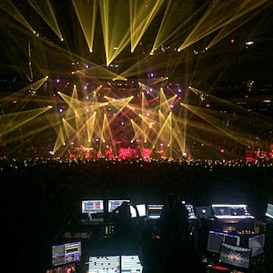 May include: A concert stage with a large crowd in front of it. The stage is lit with yellow and purple spotlights. There are several people working at a soundboard in the foreground.