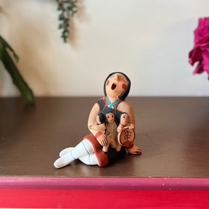 May include: A hand-painted ceramic figurine of a Native American woman holding two children. The woman is wearing a brown and white dress with turquoise accents. The children are wearing black and white dresses. The figurine is sitting on a wooden surface.
