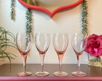 May include: Four clear pink wine glasses with a stem and a round bowl. The glasses are arranged in a row on a brown surface.