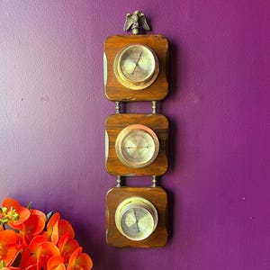 May include: A vintage wooden weather station with three circular gauges. The wooden frame is a dark brown color, and the gauges have a silver-colored face with black markings. A decorative metal eagle sits atop the weather station.