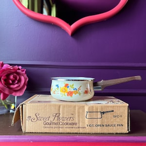 May include: A vintage cream-colored enamel saucepan with a floral design, featuring orange, yellow, and blue flowers. It has a brown wooden handle and sits on a cardboard box labeled "Sweet Flowers Gourmet Cookware". The box indicates it's a 1-quart open sauce pan.