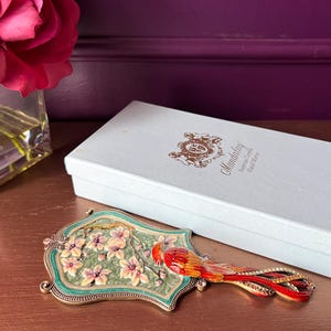 May include: A decorative hand mirror with a bird design. The mirror features a green and gold frame, a floral design, and a red and orange bird. A white box with gold lettering is behind the mirror.