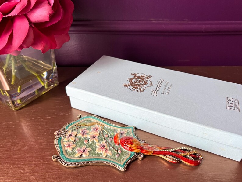 May include: A decorative hand mirror with a bird design. The mirror features a green and gold frame, a floral design, and a red and orange bird. A white box with gold lettering is behind the mirror.