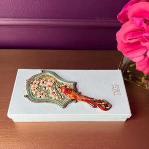 May include: A decorative hand mirror with a floral design and a red bird on top of a white box. The mirror has a green and gold frame. The box has the text "TWOS COMPANY". A pink rose is in a glass vase.