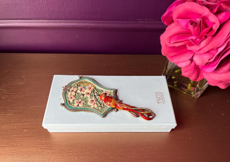 May include: A decorative hand mirror with a floral design and a red bird on top of a white box. The mirror has a green and gold frame. The box has the text "TWOS COMPANY". A pink rose is in a glass vase.