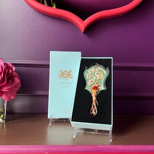 May include: A decorative hand mirror with a bird design, presented in a light blue box. The mirror features a colorful bird with a long tail, set against a floral background. The box has the word "Mandalay" printed on it.