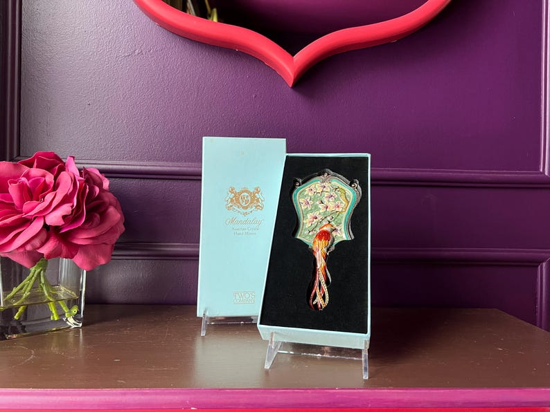 May include: A decorative hand mirror with a bird design, presented in a light blue box. The mirror features a colorful bird with a long tail, set against a floral background. The box has the word "Mandalay" printed on it.