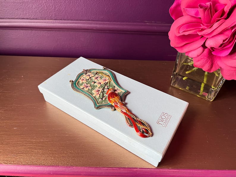 May include: A white box with a decorative bird-shaped mirror on top. The mirror has a green and gold frame with a floral design and a red and orange bird. The box has the text "TWO'S COMPANY".