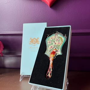 May include: A decorative hand mirror with a bird design, presented in a light blue box. The mirror features a colorful bird with a long tail, set against a floral and teal background. The box has gold lettering.