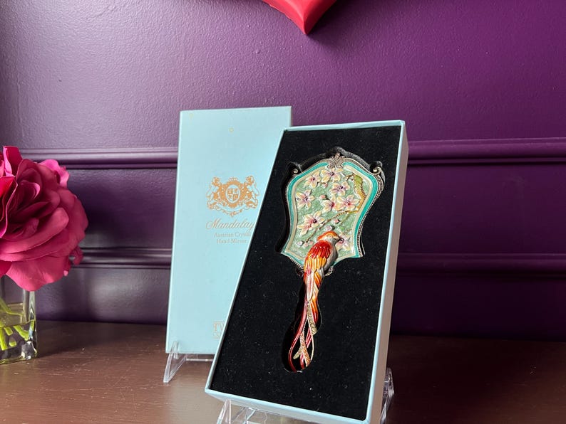 May include: A decorative hand mirror with a bird design, presented in a light blue box. The mirror features a colorful bird with a long tail, set against a floral and teal background. The box has gold lettering.