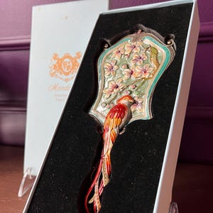 May include: An ornate hand mirror with a colorful parrot design. The mirror features a decorative frame with a floral and bird motif, presented in a gift box. The parrot has red, orange, and yellow feathers.