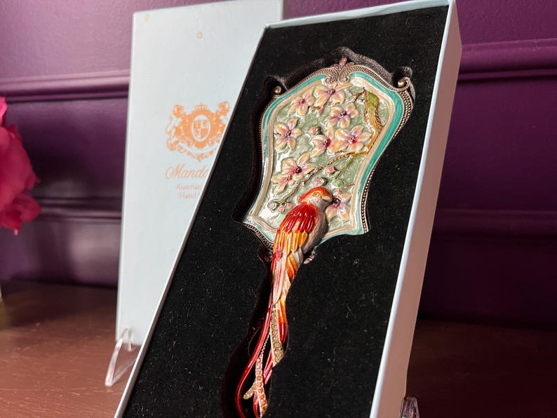 May include: An ornate hand mirror with a colorful parrot design. The mirror features a decorative frame with a floral and bird motif, presented in a gift box. The parrot has red, orange, and yellow feathers.