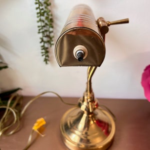 Może przedstawiać: Złota lampa biurkowa z matowym kloszem ze szkła i czarnym włącznikiem/wyłącznikiem. Lampa ma giętki ramion i okrągłą podstawę.