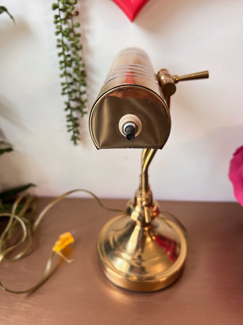 Może przedstawiać: Złota lampa biurkowa z matowym kloszem ze szkła i czarnym włącznikiem/wyłącznikiem. Lampa ma giętki ramion i okrągłą podstawę.