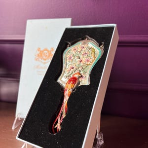 May include: An ornate hand mirror with a bird design, presented in a white box. The mirror features a colorful bird with red, orange, and yellow feathers. The mirror's frame is decorated with flowers and a light green border.
