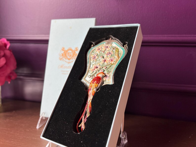 May include: An ornate hand mirror with a bird design, presented in a white box. The mirror features a colorful bird with red, orange, and yellow feathers. The mirror's frame is decorated with flowers and a light green border.