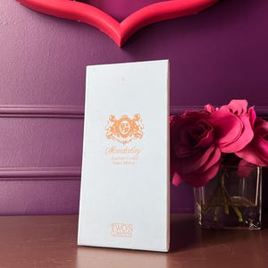 May include: A white box with the text "Mandalay Austrian Crystal Hand Made" in orange script and a decorative emblem. A bouquet of pink roses in a square glass vase sits to the right. The background is a purple wall.