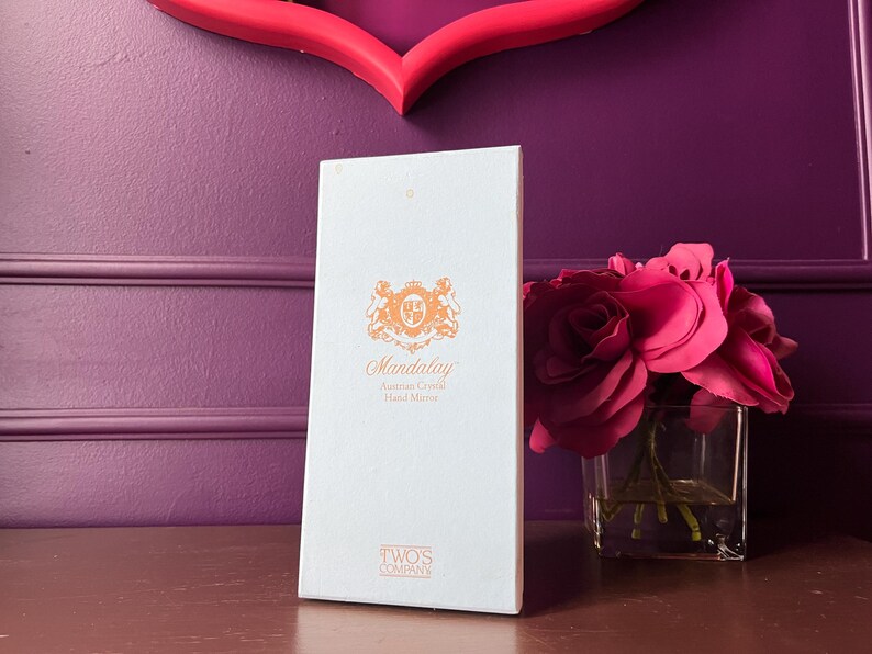 May include: A white box with the text "Mandalay Austrian Crystal Hand Made" in orange script and a decorative emblem. A bouquet of pink roses in a square glass vase sits to the right. The background is a purple wall.
