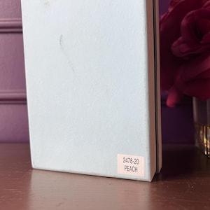 May include: A light blue box with a pink label that reads "2478-20 PEACH." The box is on a wooden surface, with a blurred background featuring a vase of red roses.