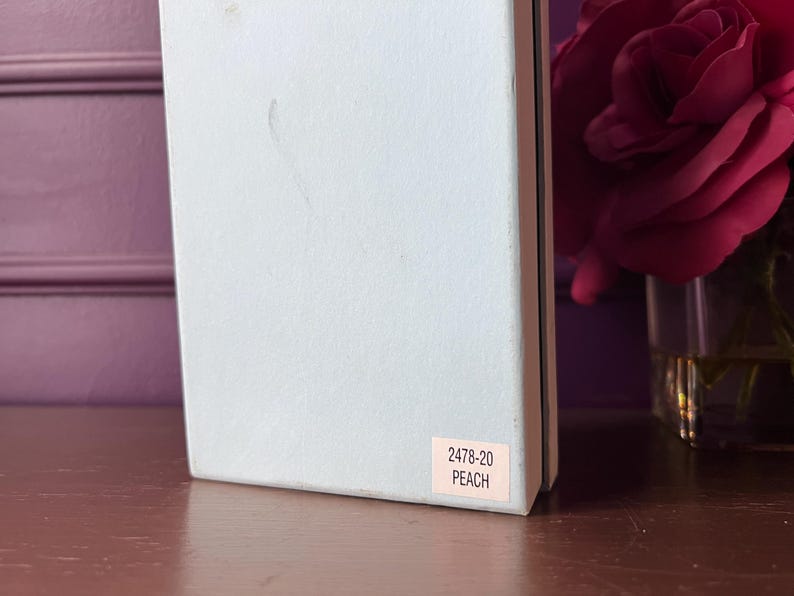 May include: A light blue box with a pink label that reads "2478-20 PEACH." The box is on a wooden surface, with a blurred background featuring a vase of red roses.