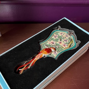 May include: Decorative hand mirror with a bird design. The mirror features a colorful bird with red, orange, and yellow feathers, perched on a floral background. The mirror is set in a teal and silver frame, presented in a light blue box with a black interior.