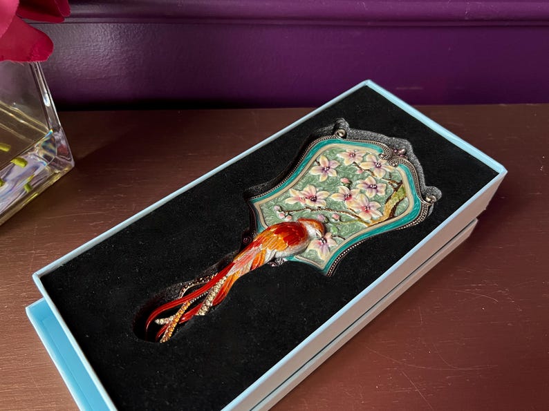 May include: Decorative hand mirror with a bird design. The mirror features a colorful bird with red, orange, and yellow feathers, perched on a floral background. The mirror is set in a teal and silver frame, presented in a light blue box with a black interior.