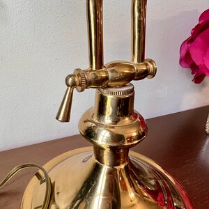 Może przedstawiać: Złota lampa biurkowa z okrągłą podstawą i jednym ramieniem. Lampa ma dźwignię na ramieniu do regulacji światła.
