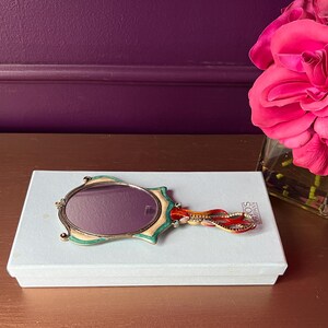 May include: An oval-shaped hand mirror with a decorative handle rests on a white box. The mirror has a gold-colored frame with green and cream accents. The handle features red and gold embellishments. A bouquet of pink roses is in the background.