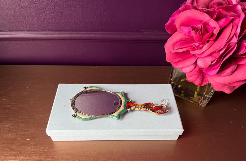 May include: An oval-shaped hand mirror with a decorative handle rests on a white box. The mirror has a gold-colored frame with green and cream accents. The handle features red and gold embellishments. A bouquet of pink roses is in the background.