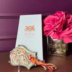 May include: An ornate hand mirror with a bird design, featuring red, orange, and yellow accents, rests on a table. The mirror is displayed with a matching box and a bouquet of pink roses in a glass vase. The mirror is approximately 6 inches long.