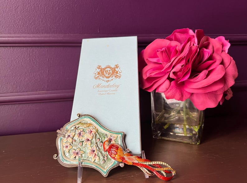 May include: An ornate hand mirror with a bird design, featuring red, orange, and yellow accents, rests on a table. The mirror is displayed with a matching box and a bouquet of pink roses in a glass vase. The mirror is approximately 6 inches long.