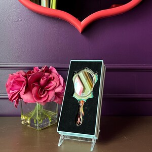 May include: An ornate hand mirror in a display box. The mirror has a decorative handle and frame with a small, round reflective surface. The box is light blue and the mirror is set against a black velvet background. A bouquet of pink roses sits nearby.