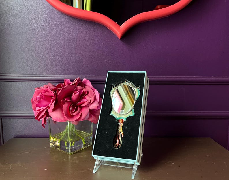 May include: An ornate hand mirror in a display box. The mirror has a decorative handle and frame with a small, round reflective surface. The box is light blue and the mirror is set against a black velvet background. A bouquet of pink roses sits nearby.
