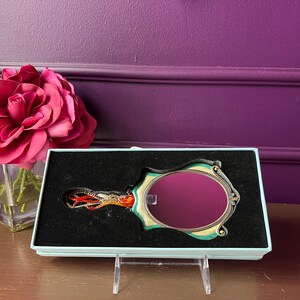 May include: A decorative hand mirror with an oval-shaped reflective surface, set in a teal and gold frame. The handle features an ornate design with red and orange accents. The mirror is presented in a light blue box.
