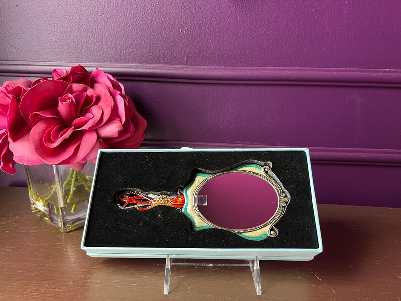 May include: A decorative hand mirror with an oval-shaped reflective surface, set in a teal and gold frame. The handle features an ornate design with red and orange accents. The mirror is presented in a light blue box.