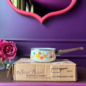 May include: A vintage 1-quart open sauce pan with a floral design in yellow, orange, and blue. The pan has a brown handle and sits on a cardboard box labeled "Sweet Flowers Gourmet Cookware."