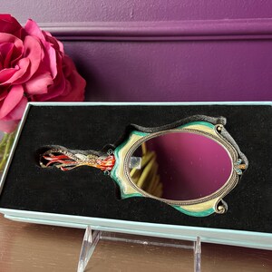 May include: An ornate hand mirror with a teal and gold frame, set in a black velvet-lined box. The mirror has an oval shape and is decorated with small jewels. A large pink rose is in the background.