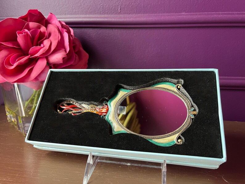 May include: An ornate hand mirror with a teal and gold frame, set in a black velvet-lined box. The mirror has an oval shape and is decorated with small jewels. A large pink rose is in the background.