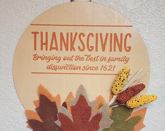 May include: A round wooden Thanksgiving sign with the word "THANKSGIVING" in orange lettering. The sign includes the phrase "Bringing out the best in family dysfunction since 1621." It is decorated with burlap leaves and decorative corn.