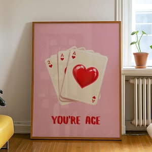 May include: A framed art print featuring three playing cards: the Ace of Hearts and the Ace of Diamonds, and the Two of Hearts, set against a pink background. The phrase "YOU'RE ACE" is printed in red below the cards.