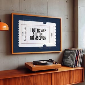 May include: A framed print with the text "I BET U2 ARE SHITTIN' THEMSELVES" in a ticket design. The print is in a wooden frame, above a record player and vinyl records on a wooden cabinet.