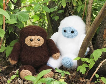 May include: Two crocheted stuffed animals, one brown and one white, are sitting in a bed of green leaves. The brown animal has a brown face with black eyes and a brown mouth. The white animal has a blue face with black eyes and a blue mouth.