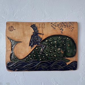 May include: A rectangular ceramic tile featuring a whimsical scene. A dark-clad figure with a crown and trident rides a green whale over blue waves. The background is tan with a stylized tree and a circular emblem.