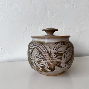 Könnte beinhalten: Ein kleiner, runder Keramiktopf mit Deckel. Der Topf ist cremefarben mit braunen abstrakten Designs und einem kleinen, abgerundeten Griff auf dem Deckel. Der Topf steht auf einer weißen Oberfläche.