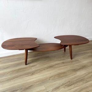 May include: A mid-century modern coffee table with a unique design. The table is made of dark brown wood and features three connected sections. The two outer sections have a curved shape, and the middle section is circular. The table is supported by three tapered legs.