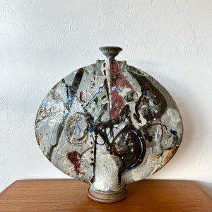 Puede incluir: Un jarrón grande de cerámica blanca con una base redonda y plana y un diseño abstracto único. El jarrón está decorado con salpicaduras de pintura marrón, rosa, verde y azul.