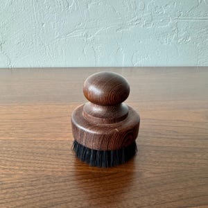 May include: A wooden brush with a dark brown finish. The brush has a rounded top and a circular base with black bristles. The wood grain is visible on the surface. The brush is designed for cleaning.