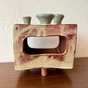 May include: A handmade ceramic candle holder with a rectangular shape and a cutout in the center. The holder is primarily reddish-brown with a light green trim and three small, light green candle holders on top. It has a single, small foot.
