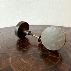 May include: Antique bronze door knob set with a round, aged patina. The set includes two circular knobs connected by a threaded rod. The knobs have a diameter of approximately 2.5 inches.