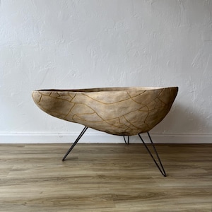 May include: A large, light brown, oval-shaped planter with a cracked, textured surface and dark brown metal hairpin legs. The planter is empty and sits on a wooden floor against a white wall.